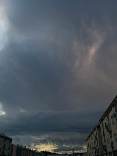The image shows a dusky sky filled with dark, heavy clouds over a residential area in ЖК «Борисоглебское», Russia. The sky occupies the majority of the frame, with the tops of buildings visible along the bottom. The clouds appear to be moving, with some lighter patches suggesting the presence of the setting sun or breaks in the cloud cover. The buildings are multi-story structures with visible windows and balconies. The overall mood is somber, with the dark clouds creating a sense of impending weather.