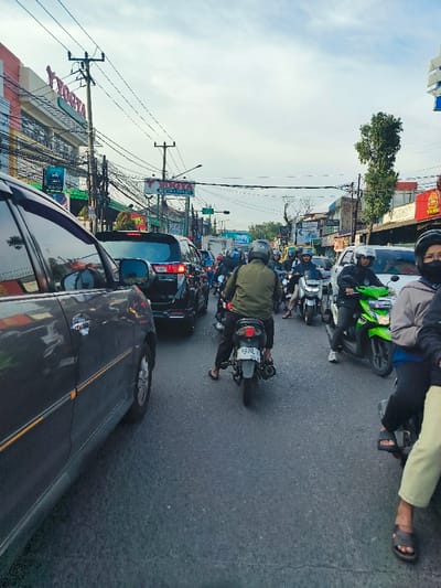 The scene is a street in Bojongsoang, Indonesia, captured at latitude -6.969 and longitude 107.637. Several vehicles are stopped in traffic, including a gray minivan, a dark SUV, and multiple motorcycles. The gray minivan is closest to the viewer and is partially obscured by the right edge of the frame. The driver of the minivan is not visible. The dark SUV is in front of the minivan. The driver of the SUV is not visible. There is a person in the driver's seat of a motorcycle. The motorcycle is directly in front of the SUV. The motorcycle driver is wearing a green shirt, helmet, and gloves. Behind the motorcycle is a second motorcycle with a driver. The driver of the motorcycle is wearing a red helmet. The scene is typical of a busy street in Bojongsoang, Indonesia. The street is paved and there are buildings along the sides of the street. The sky is cloudy. The weather is clear.