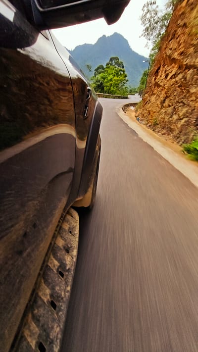 A low-angle, blurred photograph, taken from within a moving vehicle, shows a winding road in Cán Tỷ Commune, Vietnam. In the foreground, the side of a dark-colored vehicle is visible, featuring a textured side step and a partial view of the front wheel well. The road, predominantly gray with linear motion blur, stretches into the distance, curving around a rock face to the right. The rock face is rough, textured, and varies in shades of brown. In the middle distance, the road reveals a curve with green foliage and a guardrail. Further in the background, a mountain range covered in green vegetation is visible under a cloudy sky. A utility pole can be seen next to the road.
