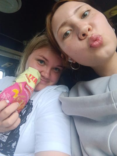 Two young women are taking a selfie in what appears to be a vehicle. The woman in the foreground on the left is holding a bag of snacks and smiling. She is wearing a white t-shirt with black lace trim. The woman on the right is puckering her lips as if to kiss the camera. She has green eyes and is wearing a grey hoodie. The background is dark and out of focus, suggesting the photo was taken indoors. The location is not identifiable from the image itself.