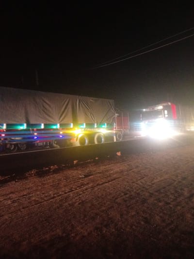 The image depicts a nighttime scene on a dirt road, possibly in Tundun Wada South, Nigeria. Two large trucks are the primary subjects. The truck on the left is a trailer with a large, dark tarp covering its cargo. Underneath the tarp, colorful LED lights (green, blue, and red) are visible along the side of the trailer, illuminating the lower portion of the vehicle. The truck on the right is a semi-trailer truck, its front cab illuminated by extremely bright headlights, obscuring details of the cab itself. A partial red section is visible on the top right of the cab. The road surface appears to be rough and muddy, with tire tracks visible. The overall impression is of a late-night road or transport activity. There are overhead wires visible in the dark sky, suggesting the trucks are on or near a road with some infrastructure. The lighting from the trucks creates a stark contrast with the dark surroundings, and the bright headlights on the right-hand truck cast a strong glow. The ground is illuminated by ambient light and reflections from the truck lights, revealing a reddish-brown, earthy texture. No people are visible in the image. The time of day is clearly night, and the weather appears to be clear.