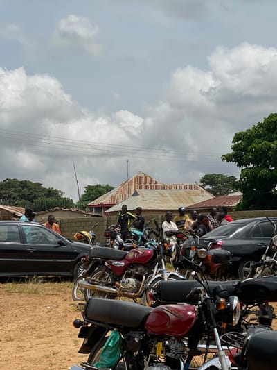 The image depicts a street scene in Kassa, Nigeria, under a cloudy sky. A group of men are gathered around several motorcycles and two black cars parked on a dirt road. The men, dressed in casual attire, are standing and talking. The motorcycles are positioned in the foreground and middle ground, with some of the men sitting on them. In the background, there are buildings with corrugated metal roofs and a few trees. Power lines are visible across the sky. The overall impression is of a casual gathering in a semi-urban environment.