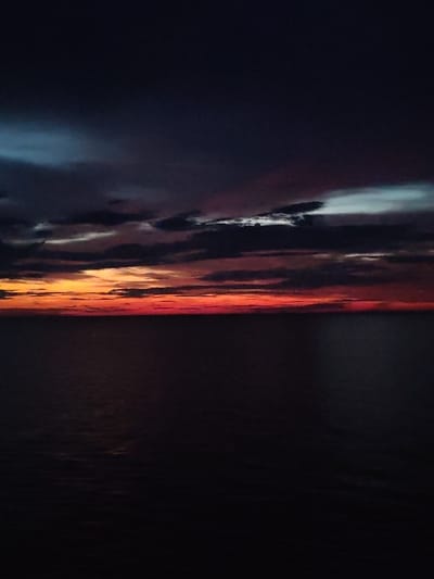 The image depicts a dark, cloudy sky over a body of water, likely the sea, at dusk or dawn. The horizon is low and illuminated by a band of orange and red light, suggesting a sunset or sunrise. The clouds are dark and striated, with hints of blue and white in some areas. The water is dark and appears calm, reflecting the dim light from the sky. There are no people, structures, or man-made objects visible in the image. The location in Kvartsitnyi, Russia, cannot be determined from the image itself, as it lacks any specific geographical markers.