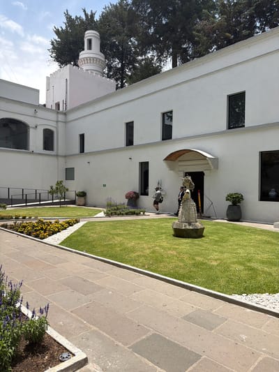 This image captures a bright, sunny outdoor scene featuring a white, multi-story building and a well-maintained garden area. The architecture of the building combines clean lines with a distinctive round, multi-tiered tower structure visible on the left side, topped with circular detailing, suggesting a blend of modern and traditional styles. Dark-framed rectangular windows are interspersed across the white facade.
In the foreground, a neatly manicured green lawn is bordered by a paved stone path composed of large, rectangular blocks. White gravel accents outline parts of the lawn and flowerbeds, which contain both purple (possibly lavender) and bright yellow flowering plants. Large, mature trees with dense dark green foliage flank the left side of the building, providing a natural backdrop.
Near the building's main arched entrance, a striking, intricately detailed statue stands on a dark, geometric pedestal. The statue depicts a figure, possibly in ceremonial attire or armor, made of a textured, light-colored material. Two people are visible near the statue: one partially obscured behind it, and another further to the left, wearing a light-colored top and shorts, looking towards the statue. They appear to be walking through the space. Potted plants and other decorative elements are placed around the building's entrance and along the garden borders.
The sky is a clear light blue with a few scattered white clouds, indicating good weather and bright daylight conditions. No discernible text is visible on any signs or objects within the frame. The overall impression is one of a meticulously cared-for architectural and garden space, possibly within a private estate or a public attraction in San Lucas el Grande, Mexico.