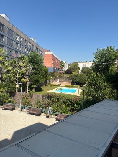 This image depicts a residential courtyard or garden area in Gavà, Spain, bathed in bright sunlight. The scene features a bright blue swimming pool surrounded by lush green grass and an abundance of vibrant green foliage, including trees and flowering bushes with pink blossoms. Several wooden benches are arranged along a paved pathway, providing seating for relaxation.

In the background, various residential buildings are visible. A prominent, modern-looking building with numerous balconies and windows dominates the left side of the frame. To the right and further back, an older, red brick building with a distinctive architectural style can be seen. Other, lighter-colored buildings are also present, suggesting a densely populated urban or suburban environment. The sky is a clear, cloudless blue, indicating fair weather.

The composition suggests a leisurely daytime scene, likely during the warmer months, given the presence of the swimming pool and abundant greenery. The overall impression is one of a well-maintained, tranquil outdoor space within a residential complex. There is no visible text in the image.