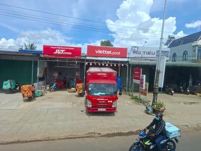 In the city of Đại Ninh, Vietnam, a scene unfolds on a street with shops and a delivery service center. A bright red Viettel Post delivery truck, an Isuzu model with the license plate "29KT-201.40", is parked in front of a Viettel Post storefront. The truck has the Viettel Post logo and the text "ISUZU 05-LOG" on its front. Adjacent to the Viettel Post shop is a J&T Express storefront. Several motorcycles are parked in the area, some equipped with delivery bags, suggesting a busy logistics hub. One motorcycle, a blue Suzuki, is ridden by a man wearing a dark jacket, blue jeans, a helmet, and a face mask, carrying a light blue storage container on the rear. In the background, other buildings and shops are visible, including one with a sign that reads "HỦ TIẾU 1994". The sky is partly cloudy, indicating daytime. The ground is paved with concrete tiles.