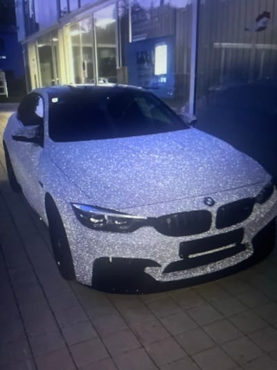 A highly reflective BMW car, covered in glitter, is parked on a paved surface outside a modern building with large glass windows. The car has its headlights on, illuminating the front grille and bumper. The building appears to be a commercial or residential property. The scene is likely in an urban setting, possibly a street or parking lot in the city of Kołobrzeg, Poland. No people are visible in the image.