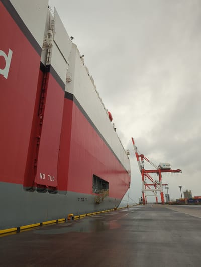 This is a wide-angle, low-angle shot of a large red and white ship docked at a port in Chiba, Japan, on an overcast day. The ship occupies the left two-thirds of the frame, its massive hull painted red and gray. "NO TUG" is stenciled in white letters on a red section of the ship's hull. To the right of the ship, a large red and white gantry crane stands tall against the sky, with another, larger crane partially visible behind it. In the distance, more cranes and stacks of shipping containers can be seen. The ground is wet and reflective, suggesting recent rain. The scene is calm and industrial, with no people or visible activity.
