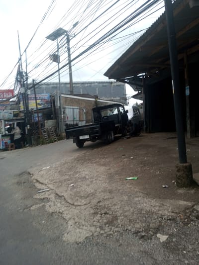 The image shows a street scene in Ban Phouviengnoy, Laos. A dark-colored utility truck is parked on a dirt and paved road surface. A person appears to be working on the truck's wheel, positioned near a building with a corrugated metal roof and open front. Across the street, there's a storefront with advertising signage and people standing near it. Above, a dense network of overhead wires and poles crisscrosses the sky. The general environment appears gritty, with litter scattered on the ground. The sky is overcast.
