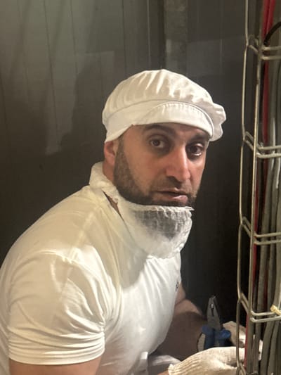 In a close-up, medium-shot, a man wearing a white cap and a white shirt with a disposable beard net over his lower face looks into the camera. The man has dark hair and a beard and is looking slightly to his left. To the right of the man, a bundle of cables is visible, some of which are gray and red. A pair of pliers with blue handles rests on a white cloth in the foreground, and the man's arm, also wearing a white cloth, is visible. The background consists of a dark, textured wall.