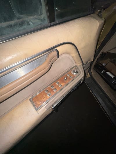 The scene captures the interior of a vehicle, specifically a front door panel, located in Zhabkino, Russia. The door panel is upholstered in worn, light-tan padded material, exhibiting creases and dirt accumulation. A horizontal chrome trim piece is present above a recessed armrest section, which includes a matching tan door pull handle. Below the armrest, a control module with a distressed faux woodgrain finish houses four silver-colored rectangular power window switches, arranged in two columns of two. To the right of these switches are two circular controls, one larger and one smaller, for unspecified functions. The lower portion of the image shows a dark floorboard inside the vehicle and dark ground outside the open door. Indistinct dark objects are visible on the vehicle's floorboard to the far right. A dirty vehicle window is partially visible in the upper left. No individuals are present.