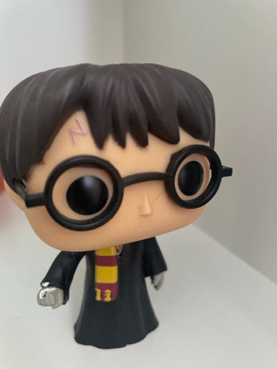The media file shows a close-up, rotated view of a Harry Potter Funko Pop figurine, likely displayed indoors. The figurine features the iconic character with dark, molded hair, round black glasses, and a pale complexion. A pink lightning bolt scar is visible on its cheek. The figurine is wearing a black robe with a red and yellow striped scarf. One of its hands is clenched into a fist, painted a light gray with white paint streaks. The background is a plain, light-colored surface, possibly a wall or shelf, common in indoor settings. No specific time of day, weather, or explicit location cues are discernible, other than the provided location context of Otradnoye, Russia, which is external information. There is no visible text within the image itself. The overall presentation suggests a still image of a collectible toy.