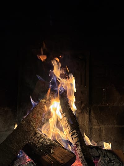 A fire burns within a stone or brick fireplace, located in Huasca de Ocampo, Mexico. Several wooden logs, exhibiting charring and visible wood grain, are stacked in the hearth. Glowing red and orange embers are present at the base of the logs. Vibrant yellow and orange flames, with areas of blue at their core and edges, emanate from the burning wood, rising upward. Faint gray smoke is visible above the flames. The interior walls of the fireplace, particularly on the right, are constructed of dark, possibly sooty, brickwork. No individuals, additional objects, or external light sources are present in the immediate scene; the fire itself provides all illumination, contrasting against the dark backdrop.
