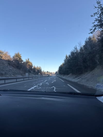 The image captures a forward-facing view from the interior of a vehicle, looking through a dirty windshield with visible streaks and smudges. The scene depicts a multi-lane paved road, likely a highway or major route, extending into the distance under a clear, light blue sky. Irregular white patterns, consistent with de-icing salt residue or dried liquid, are present on the dark asphalt road surface, primarily in the right lane.

To the left of the road, a silver metal guardrail runs parallel to the vehicle's direction of travel. Beyond the guardrail, an embankment rises, covered with a mixture of evergreen and bare deciduous trees, and ground vegetation showing white frost. To the right of the road, a similar frosted embankment slopes upwards, densely forested with evergreen trees, also displaying frost on their branches and the ground. The overall environment suggests cold, clear weather conditions. No individuals are visible. The location is in Monistrol-sur-Loire, France.
