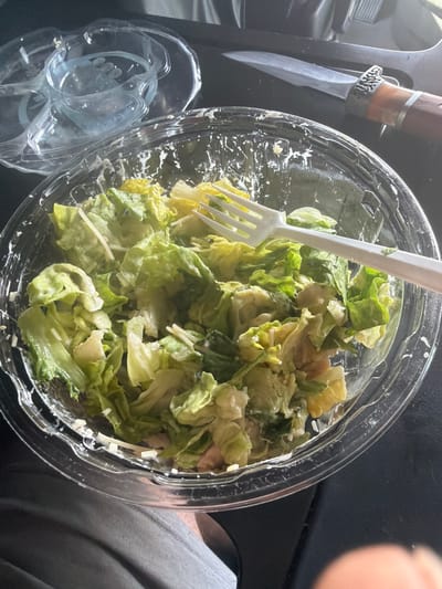 A plastic salad bowl filled with lettuce and dressing sits on a dark surface, possibly a car interior, with a white plastic fork resting on top. To the left, a stack of clear plastic containers is visible, and to the right, a knife with a wooden handle and a metal blade is positioned next to the salad. The overall scene suggests a meal is being consumed or prepared.