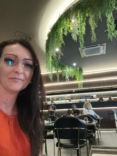 The image is a selfie taken in a restaurant with a contemporary design. The foreground features a woman wearing glasses and an orange top, with her dark hair falling over her shoulders. In the background, several people are seated at tables. One man is standing, possibly addressing the diners. The restaurant's interior is decorated with hanging greenery and features a modern lighting system. The overall atmosphere suggests a casual dining experience. The scene is located in Ponteterra, Italy.