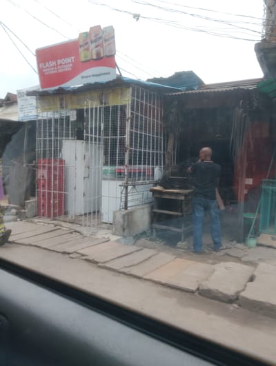 The image depicts a street scene in Alagbede, Nigeria. In the foreground, a man with a shaved head, wearing a black t-shirt and blue jeans, stands facing away from the camera, appearing to be engaged in work or conversation. He is positioned in front of a shop or stall.

The shop itself is open-fronted and appears to be a business that sells various items. There are metal cages or grills protecting the interior, and inside, an array of goods are visible, including what look like coiled cables or ropes hanging from the ceiling and stacked on shelves. There are also some large, white appliances that could be refrigerators or freezers visible behind the grills. To the left of the shop, there is a red-doored appliance, possibly another refrigerator, also behind a protective grill.

Above the shop, a prominent red and white sign announces "FLASH POINT INDOOR & OUTDOOR CATERING SERVICES" and features images of Maltina beverage cans. Below this, smaller text reads "Share Happiness." Further to the left, a partially obscured sign with blue lettering reads "Ola-Plus."

The ground in front of the shop is paved with a mixture of wooden planks and concrete slabs. The overall setting suggests a busy commercial area, likely in a developing urban or semi-urban environment. The lighting indicates it is daytime, but the sky appears overcast, suggesting potentially cloudy weather. The scene is typical of a street-side market or service provider in Nigeria.