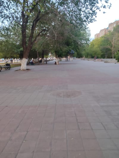 The image depicts a paved park area with trees lining a pathway. A multi-story building, typical of Soviet-era architecture, is visible in the background on the right. Several individuals are present in the park. On the left side, a person is riding a bicycle, and further down the path, another individual appears to be on a bicycle or scooter. Closer to the center, two people are seated on a low stone wall. The ground is paved with interlocking square tiles, with a distinct reddish-brown hue in the foreground. Trees with white-painted bases are scattered throughout the park. The sky is overcast, suggesting it is either early morning or late afternoon. The scene is consistent with public parks in residential areas of Temirtau, Kazakhstan.