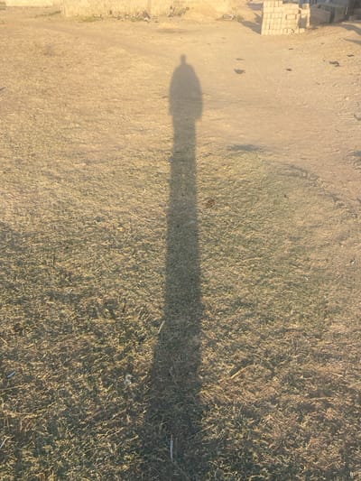 The image depicts a shadow figure cast on a mixed terrain of dry dirt and sparse grass. The shadow is elongated and appears to be that of a person or a small structure with a rounded top. Adjacent to the dirt area is a pile of bricks or paving stones. The scene is bathed in warm sunlight, contributing to the stark contrast between the shadow and the surrounding ground.