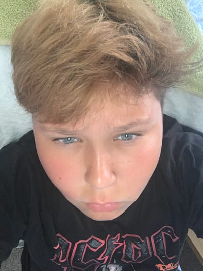 A close-up, rotated image shows the face of a person with light brown hair and blue eyes. The person is wearing a black t-shirt with a graphic design featuring red and black lettering that appears to be text. The background is blurry, but suggests an indoor setting with a textured green surface and a white surface. The lighting suggests it could be daytime, possibly indoors. The person's expression is neutral. The graphic on the t-shirt is partially visible and features bold, stylized lettering. The image is a selfie, taken from a high angle, focusing on the person's face. The location is Vejprty, Czechia.
