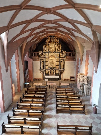 This high-angle photograph depicts the interior of the Collegiate Church of St. Peter and Alexander in Aschaffenburg, Germany, viewed from the rear gallery looking towards the altar. The scene is devoid of people.
The ceiling features a prominent ribbed vaulting system composed of light plaster infill and robust reddish-brown sandstone ribs forming an intricate pattern. The side walls are predominantly light-colored stucco, contrasting with vertical reddish-brown pilasters and arch structures. The floor is tiled with a checkerboard pattern of alternating light beige and reddish-brown square tiles.
The focal point is a large, highly elaborate, multi-tiered wooden altarpiece (reredos) at the far eastern end. It is richly carved with numerous figures, architectural elements, and narrative scenes, appearing in dark, possibly ebonized wood, with lighter, possibly gilded, carved sections. A simple white altar cloth drapes a table in front of the reredos, flanked by two candles. To the right, an ornate carved stone pulpit with multiple sculptural reliefs is attached to the wall.
The nave is furnished with two symmetrical blocks of dark wooden pews, each featuring light-colored seat pads or cushions. These pews are arranged in rows, creating a central aisle leading directly to the altar. In the foreground and mid-ground, several dark metal display tables, some holding informational materials, are positioned along the side aisles. On the upper left, above the altar area, a wooden gallery with a dark balustrade is visible. A recessed arched doorway with an internal staircase is visible on the left wall further back. A tall, cylindrical, dark-colored container with a metallic basin top stands against the far right wall. The space is illuminated by a combination of diffused natural light and discrete ceiling fixtures.