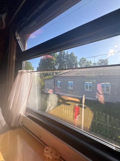 The image is taken from the interior of a room, looking up through a sunroof or window. The sky is visible through the open portion of the sunroof, and beyond the window pane, a gray brick building with multiple windows and a steeply pitched roof is visible. Trees with green foliage are positioned between the building and the window. A partial view of what seems to be an exterior of a building is visible, with a red object on it. A light-colored, patterned curtain or blind is partially drawn. The location might be Заслаўе, Belarus, based on the prompt.