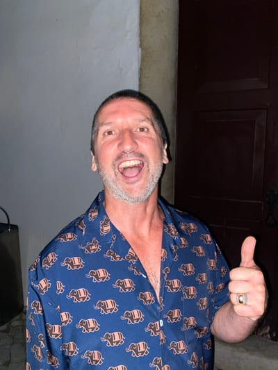 A medium shot shows a Caucasian man in his 50s wearing a dark blue short-sleeved button-up shirt with a repeating pattern of elephants in beige. He has short, graying hair, a light beard, and is smiling with his mouth open. His right arm is bent upwards, with his thumb giving a thumbs-up gesture. He is wearing a ring on his left hand. The man is standing in front of a textured white wall on his left and a dark wooden door with a decorative metal handle on his right. A thick, cylindrical stone pillar is visible between the wall and the door. The background suggests an outdoor setting, possibly in a historic part of Loulé, Portugal, given the architectural elements.