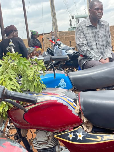 The image depicts an outdoor scene in Kassa, Nigeria, under an overcast sky. In the foreground, two motorcycles are prominently featured. The motorcycle on the left is red with colorful decals and a cluster of green leaves resting on its handlebars. The motorcycle on the right has a black seat and a blue fuel tank adorned with a sticker of a person. A man wearing a patterned button-down shirt and dark trousers is seated on the blue-tanked motorcycle, looking towards the left of the frame with a pensive expression.

Behind the motorcycles, several individuals are gathered. To the left, a woman wearing a dark top and a headscarf is partially visible. Further back, a group of women are clustered together, some wearing colorful headscarves. In the background, a brick wall and a transformer with electrical lines are discernible. The ground appears to be dusty or sandy. The overall impression is a momentary pause or gathering of people in a semi-urban or rural Nigerian setting.