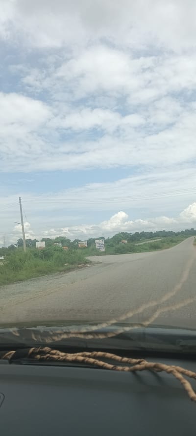The image depicts a view from inside a vehicle, looking out onto a road near Umuoye, Nigeria. The road ahead is paved and appears to curve to the right. On the left side of the road is an intersection with a smaller road or driveway. There's vegetation on both sides of the roads, with scattered buildings and trees visible in the distance. A power pole stands on the left side of the road. A sign is present near the intersection, with text on it. The sky is predominantly cloudy with patches of blue. A set of twisted, thin, rope-like objects are on the vehicle's dashboard.