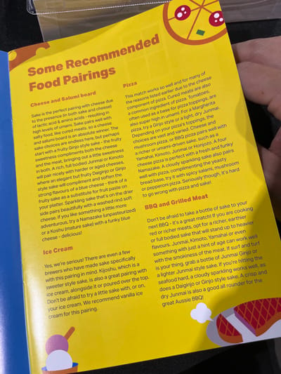 The image is a section of a printed menu board, showcasing a yellow background. The text on the board recommends specific food pairings with sake, specifically focusing on cheese and salami, pizza, BBQ and grilled meat, and ice cream. The location, based on the coordinates provided, is in the suburb of Hamilton, Brisbane, Australia. The text discusses the perfect sake pairing with cheese due to the presence of lactic acid and amino acids. It suggests trying different kinds of sake such as Junmai, Kimoto, or Daiginjo, and pairing them with various cheese options. The menu also highlights specific sake recommendations for pizza, BBQ and grilled meat, and ice cream.  The image appears to be a close-up of a menu from a restaurant or bar.