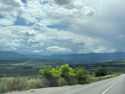 The image depicts a rural landscape near Isperihovo, Bulgaria, under a cloudy sky. In the foreground, a paved road with a white dividing line curves to the right. The roadside is bordered by dry grasses and bushes on the left and a vibrant green, leafy tree on the right.

Beyond the road, a vast expanse of green fields stretches into the distance, dotted with scattered trees and some buildings that suggest a small settlement or agricultural structures. The middle ground is dominated by rolling hills covered in lush vegetation.

In the background, several layers of mountains recede into the hazy distance, their slopes covered in green forests. The sky is filled with dramatic, textured clouds, ranging from bright white to darker grey, with patches of blue sky visible between them. The overall impression is of a vast, natural landscape under a dynamic sky. There are no people or animals visible in the image.
