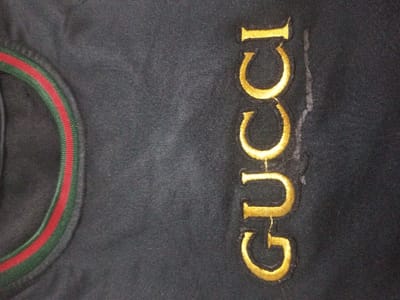The image shows a black garment with a rounded neckline trimmed with green and red stripes. Embroidered vertically on the right side of the garment is the word "GUCCI" in gold thread. The embroidery appears somewhat worn, and there is a faint, irregular stain running alongside the letters. The fabric of the garment has a faded or worn appearance.