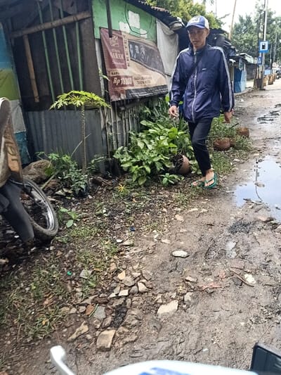 A man in a blue jacket and cap walks on a broken, muddy road next to makeshift buildings. The road is damaged and has puddles. A parked motorcycle is partially visible on the left. Vegetation grows between the road and the buildings, one of which displays an advertisement poster. A street sign can be seen in the background. The overall scene suggests a low-income, possibly informal settlement.