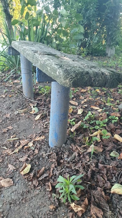 This image shows an outdoor scene featuring a weathered, concrete bench with blue, cylindrical legs. The bench is situated on uneven ground covered with fallen leaves and sparse vegetation. The background consists of foliage and trees, suggesting a park or garden setting. The overall impression is one of neglect or natural overgrowth. The location in Ust-Labinsk, Russia, is not visually indicated in the image. There are no people or animals present in the scene. The lighting suggests it is daytime.