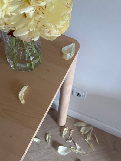 The image shows a still life composition. A light yellow flower arrangement in a clear glass vase sits on a light-colored wooden surface. The surface appears to be part of a small table or platform. Several fallen flower petals are scattered on the table and the floor, which has a light wood grain pattern. In the background, a plain white wall is visible with an electrical outlet. The angle of the shot is from above, looking down and slightly to the side. The location is unknown, and no information suggests it is in Люк, Russia.