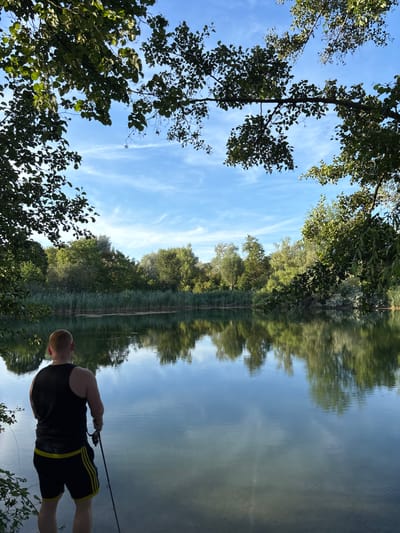 A person stands with their back to the camera, holding a fishing rod and looking out at a calm body of water. The person is wearing a black tank top and black shorts with yellow stripes. The water reflects the trees and sky, which is mostly blue with some wispy white clouds. The foreground is composed of branches and leaves from trees overhead, and the background features a dense line of green reeds and various trees. The scene appears to be a natural setting, possibly a park or lake within the city of Ludwigshafen am Rhein, Germany.
