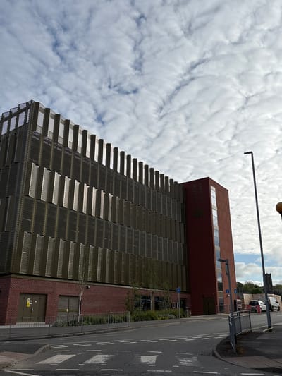 The image captures the Castle car park in Newcastle-under-Lyme, United Kingdom, from a low-angle perspective. The car park's distinctive stepped facade of a metal perforated screen dominates the foreground, adjacent to a red brick section with two closed service doors. Above this is a rust-colored, rectangular building extension with a horizontal row of windows along the top. The building has signage that is difficult to read, appearing to say "Castle Car Park."
A street runs parallel to the building. Several parked cars and vans are visible in the background. Street furniture includes a tall streetlight, pedestrian barriers, and signs.
The sky is heavily clouded, suggesting overcast weather.