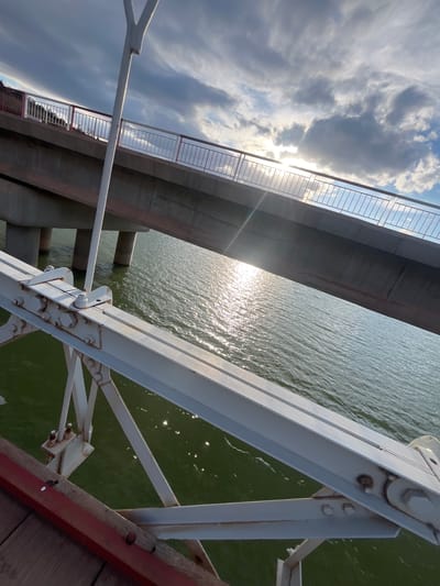 A low-angle shot depicts a bridge structure, likely in Ait Mazigh, Morocco, judging by the location request. The composition features the bridge's steel framework, painted white, in the foreground, contrasted against the backdrop of water reflecting sunlight. Another bridge structure is visible parallel to the primary bridge, identifiable by its grey concrete supports and pedestrian railing. The sky is partly cloudy, with sunlight breaking through. The water appears still, with the sun reflecting off the water. The perspective suggests the photo was taken from a point below the bridges, close to the water level.