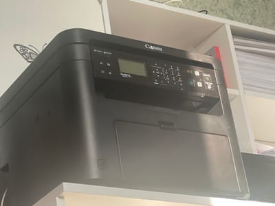 The image shows a black Canon i-SENSYS MF231 printer on a white shelf. The printer is positioned at an angle, with the top left corner closer to the viewer. A black butterfly sticker is visible on the wall to the left of the printer. The printer has a control panel with a small LCD screen, buttons for copy, scan, and other functions, and a numeric keypad. The printer is located indoors, likely in an office or home setting, with the background showing white shelving units filled with paper. The scene does not provide any information or context related to the city of Novoselskoye, Russia.