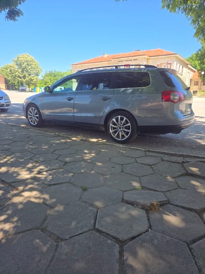 In the city of Srednia, Bulgaria, on a sunny day, a grey Volkswagen Passat wagon is parked on a sidewalk made of hexagonal paving stones. The car is positioned parallel to the curb, with its driver's side facing the camera. The vehicle's windows are tinted. In the background, a building with a terracotta roof is visible, and beyond that, trees and a bright blue sky. The sidewalk is partially shaded by the leaves of a nearby tree, casting dappled patterns of light and shadow. There are no people visible in the image.