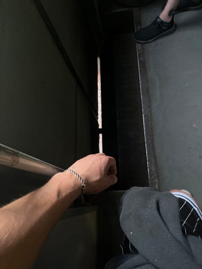 A person's left arm, adorned with a silver bracelet, rests on a metal railing in what appears to be an industrial or utilitarian setting. The arm is hairy and its skin tone is light. To the right of the arm, part of another person's leg is visible, wearing black socks and black Vans sneakers with white stripes. The background is dark and indistinct, suggesting an indoor or shadowed area. The environment seems to be a confined space with metallic textures and possibly ventilation grates. The location is identified as Hlevakha, Ukraine.