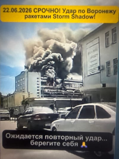 The image depicts a scene of destruction in a city, possibly Sof'ino, Russia. In the background, a building is engulfed in thick, dark smoke, suggesting a fire or explosion. Debris and smoke obscure the upper floors of the building. Several cars are visible in the foreground, parked on a street. One car, a white hatchback, is in the center of the frame, and another darker car is to its left. A black bar overlays the bottom of the image, containing text in Russian. The text appears to be an alert or warning. The overall impression is one of a recent event causing significant damage and disruption.