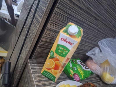 A carton of Chivita orange juice sits on a wooden surface next to a plastic bag containing a lemon and carrots, and a bag of Milo. The juice carton is mostly orange with green and white accents. The surface has a striped, wood-grain appearance. A dark, metallic object is partially visible in the upper right corner.