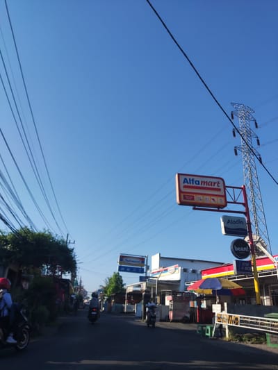 The image depicts a daytime street scene in Sidokepung, Indonesia, under a clear, bright blue sky. Numerous black electrical and communication lines crisscross the upper left and center of the frame, converging towards the left horizon. A prominent steel lattice electricity transmission tower stands on the far right, extending from the mid-ground into the sky.

The scene focuses on a paved road, likely asphalt, with multiple motorcycles in motion. On the left, a person wearing a red helmet and a light-colored top is riding a dark-colored scooter towards the viewer, partially obscured by the left edge of the frame. Further down the road in the center, another individual on a dark scooter, wearing light clothing, drives away from the viewer.

Commercial establishments line both sides of the street. On the right, a large, rectangular sign with a red border prominently displays "Alfamart" in blue lettering. Below it are smaller signs for "Aladin" and "bean! SPOT," with "ATM & BCA" visible beneath. Near these signs, a colorful, open umbrella with red, blue, and yellow panels provides shade over what appears to be a street vendor's stall, accompanied by green stools on the roadside.

On the left side of the road, dense green foliage from trees and shrubs partially obscures other buildings. Further down the street on the left, an "Indomaret" sign is visible above commercial structures. The overall environment suggests a bustling commercial and residential area with typical Indonesian street infrastructure.