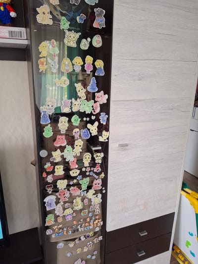 Here is a detailed description of the image:

The image shows a piece of furniture, which appears to be a cabinet or a storage unit, covered with numerous colorful stickers. The cabinet's design includes a two-toned finish: the main part is a light, faux-wood grain material, and a section to the right is a darker brown. The surface under the stickers is shiny, possibly glass.

The stickers themselves are various cartoon-like figures. There are images of animals, human-like characters, stars, and other shapes. The stickers are densely packed on the shiny surface.

In the bottom left corner of the image, there is a package labeled "MAGIC BLOCK" and colorful children's building blocks. To the left of those blocks, there is text that appears to be a product description. In the bottom right of the image, part of a black flat screen TV is visible.

There is no specific information related to the city of Ozyorny, Russia, that can be determined from the visual information present in the image. The provided OCR text states: "а Нептвенигоро Марата, д.86 Телефон +7(812)448-00-7". The phone number is a Saint Petersburg, Russia, code.