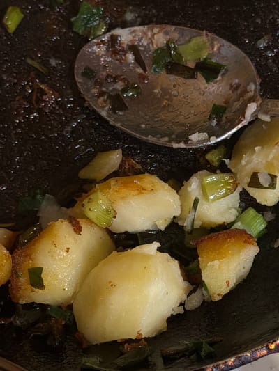 A close-up, overhead shot shows roasted potatoes with scallions in a pan, as if recently served or prepared. The pan is dark and appears to be cast iron, with some residue and browning. A silver spoon rests in the upper portion of the pan, partially scooping some of the potatoes and green onions. The potatoes are roughly cut into bite-sized pieces, some with golden-brown edges from roasting, while others remain pale yellow. The green onions are chopped into short segments and interspersed with the potatoes. The overall impression is of a rustic, home-cooked meal. This image is not associated with Satino-Russkoe, Russia, as there is no visual information to support such a location.