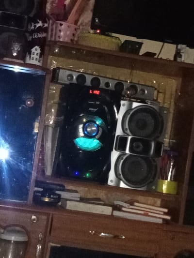 The image displays an indoor setting, likely a section of a wooden entertainment unit or shelf, showcasing various electronic devices and personal items. The overall lighting appears dim, with several electronic components emitting their own light.
At the center of the frame, there is a prominent black electronic device, possibly a subwoofer or the main unit of a sound system. Its front features a distinctive circular design with a glowing blue or cyan light, and a smaller blue LED is visible towards its base. To the right of this black unit, a silver-colored speaker column stands, equipped with two large circular speakers and a rectangular, possibly decorative or functional, element in between them. Directly above these sound components, a long, slender silver device, resembling a soundbar or an audio equalizer, is positioned. It has several circular controls or indicators along its front, and a red digital display clearly shows the word "LINE".
To the far left of the image, a large, dark, reflective surface is visible, which could be a television screen or a large mirror. A bright, starburst-like reflection of a light source is strongly apparent on its surface, indicating ambient light or a nearby lamp. Below this reflective surface, on the wooden shelf, a small black object is placed, featuring white text that reads "BLADE". A stack of books or small rectangular objects rests next to it.
In the upper left portion of the frame, parts of another dark speaker or electronic device are visible. Below it, a black mug with white holiday-themed patterns, including reindeer and a small house, is present. Partial white text on the mug suggests "Merry Christm...". Adjacent to the mug, a white, intricately cut basket can be seen.
The top right corner of the image shows a black flat-screen television, which appears to be powered on, indicated by a small red light. Below the speaker column on the right, a clear container with a yellow base holds several slender items, possibly pens or pencils. The shelving unit itself is made of wood, contributing to a warm, domestic feel. The arrangement suggests a personal space where media consumption is a regular activity.