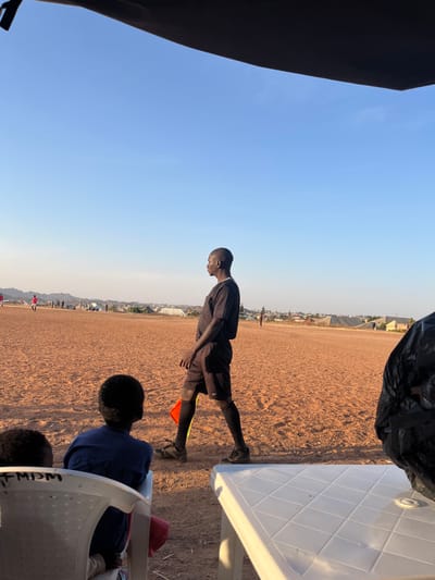 An outdoor scene in Barakin Acha, Nigeria, depicts an unpaved reddish-brown earth football field under a clear blue sky. In the mid-ground, a dark-skinned male adult, appearing to be an assistant referee, walks from left to right, carrying a red and white flag in his left hand. He is dressed in a black short-sleeved shirt, black athletic shorts, black socks, and dark athletic shoes, and looks slightly to his right. In the foreground, positioned to the left, two individuals, likely children, are seated on white plastic chairs, facing away from the camera and towards the field. The back of the visible chair has "FMPDM" inscribed on it. To the immediate right in the foreground, a portion of a white plastic table with a raised grid pattern is visible. An overhead black canopy or structure partially frames the top of the image. In the distant background on the field, several smaller figures, some in red shirts, are visible, suggesting other players. Beyond the field, a sparse urban landscape with scattered buildings and low hills is present under a bright, clear sky.