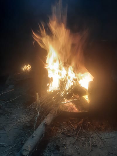 A nighttime scene depicts two fires burning in the city of Vosiobod, Tajikistan. The primary fire, larger and more intense, is contained within an open-sided structure, likely a metal brazier or kiln. Flames, ranging in color from bright yellow to orange, erupt from the burning materials, which appear to be wood and other combustible debris. The fire illuminates the immediate surroundings with a warm glow, casting shadows and highlighting the rough texture of the ground, which is covered in dirt, small stones, and scattered twigs and branches. In the background to the left, a second, smaller fire burns with less intensity, appearing to be a more modest bonfire. Both fires are generating smoke, which drifts upwards and into the dark night sky, creating a hazy atmosphere. The overall impression is one of a rural or semi-urban environment at night, where fires are being used for warmth or some other purpose.