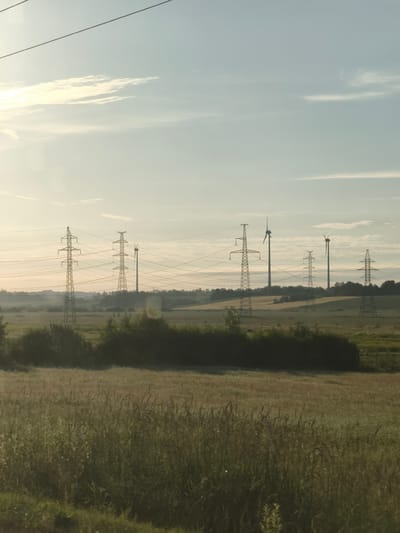 The image depicts a rural landscape with several electricity pylons and wind turbines under a hazy, pale blue sky with scattered, thin clouds. The sun appears to be low on the horizon, casting a soft light. In the foreground, a field of tall, dry grass and some greenery can be seen. Behind the grass, there is a line of dark green bushes and trees. Further in the background, the electricity pylons stand tall, connected by numerous wires. Several wind turbines are visible in the distance, positioned on rolling hills. The overall impression is one of an open, agricultural or pastoral area with infrastructure for power generation. The location is identified as Āres, Latvia.